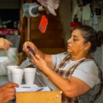 Busy food stall scene with woman serving customers, holding smartphone.
